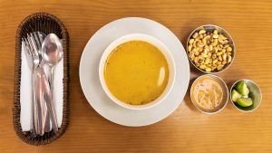 A delicious bowl of Peruvian soup with sides of corn, sauce, and lime on a wooden table.